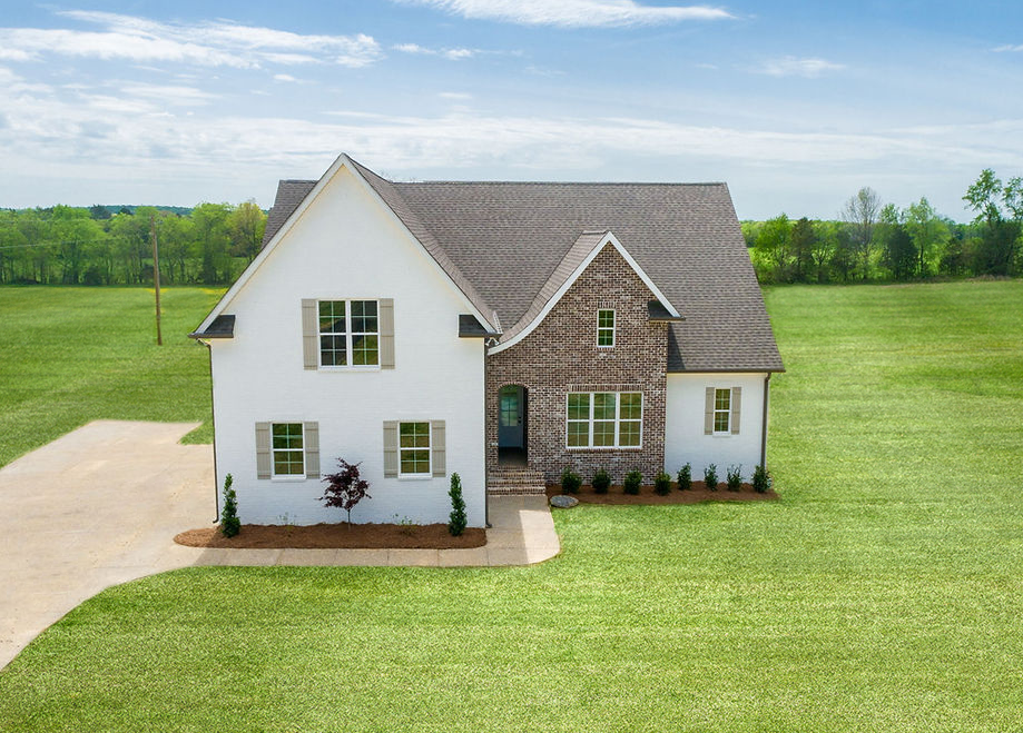 A modern farmhouse built by Stewart Knowles Construction featuring a white painted brick exterior, brown brick accent wall, gable roof, and neatly landscaped front yard surrounded by open green fields.